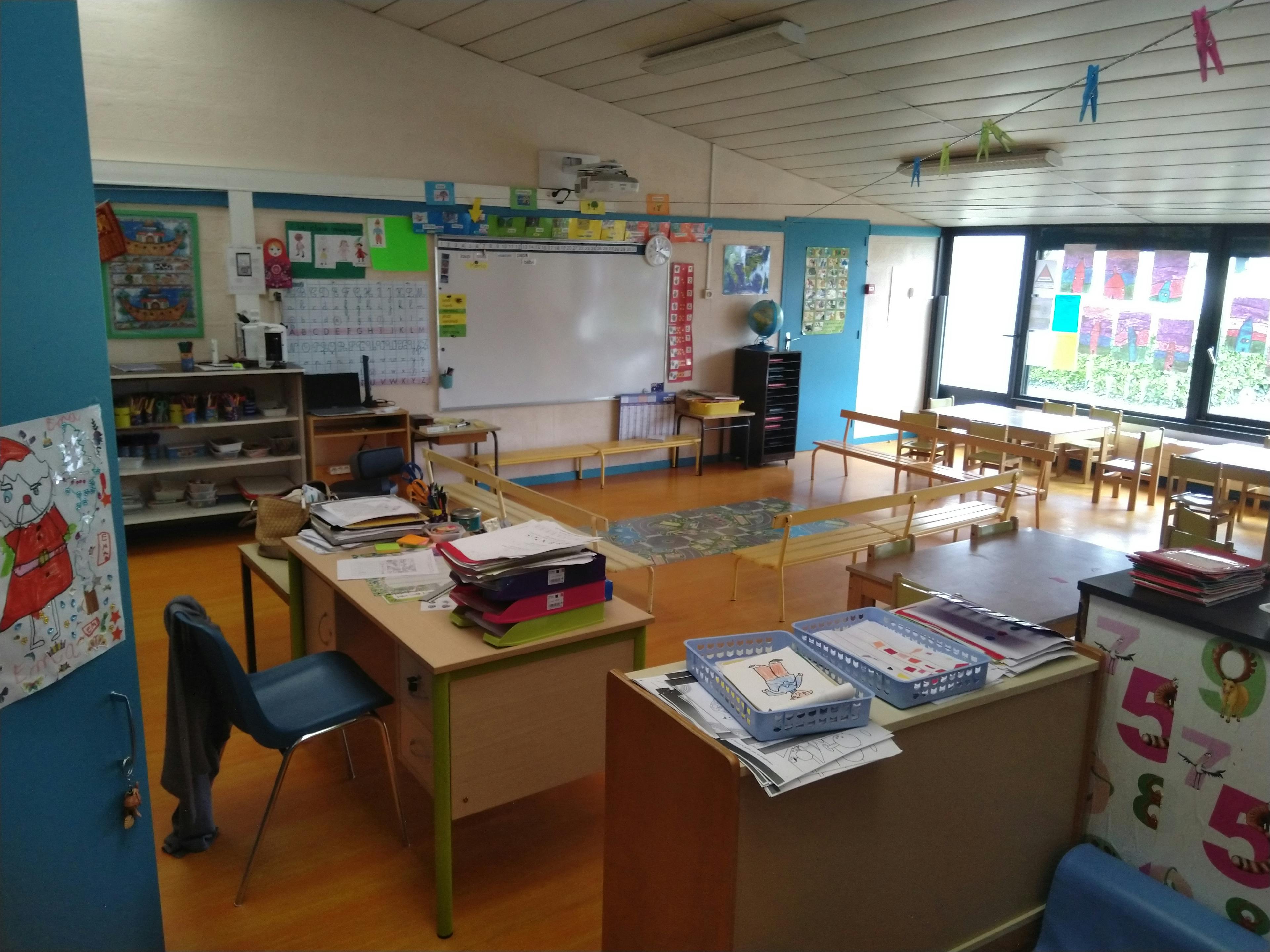 Vue intérieure d'une salle de classe avec un bureau, des fournitures rangées et des bancs.